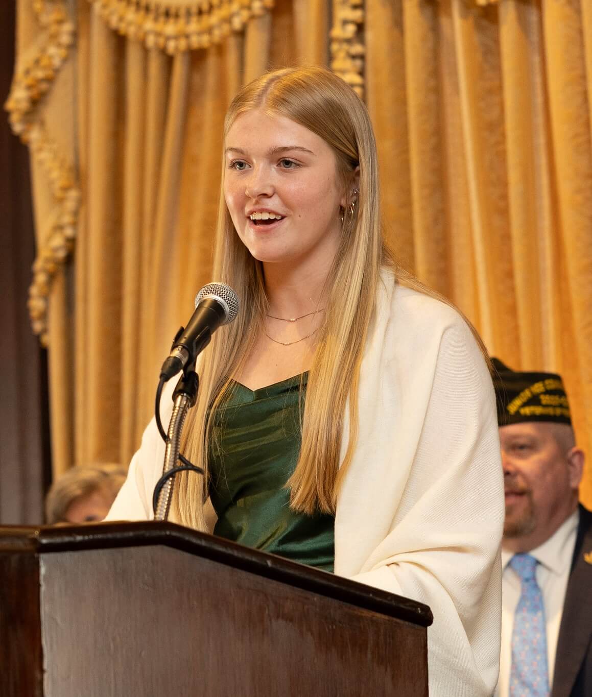 Young woman in a green dress speaks at a podium, with a microphone, in a formal setting.