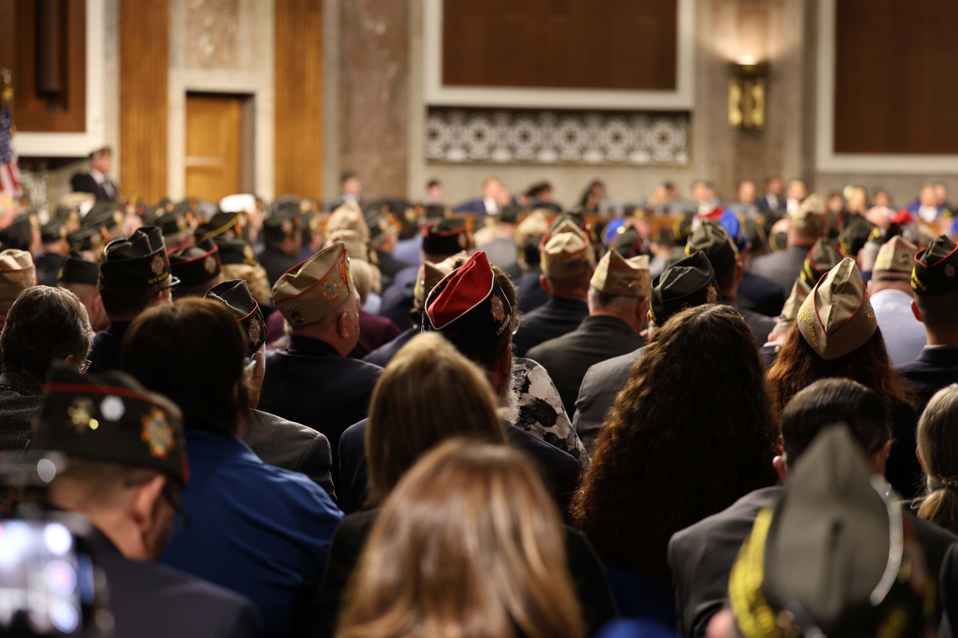 A crowd of veterans in formal attire attends a ceremony, showcasing their hats and focused attention on the proceedings.