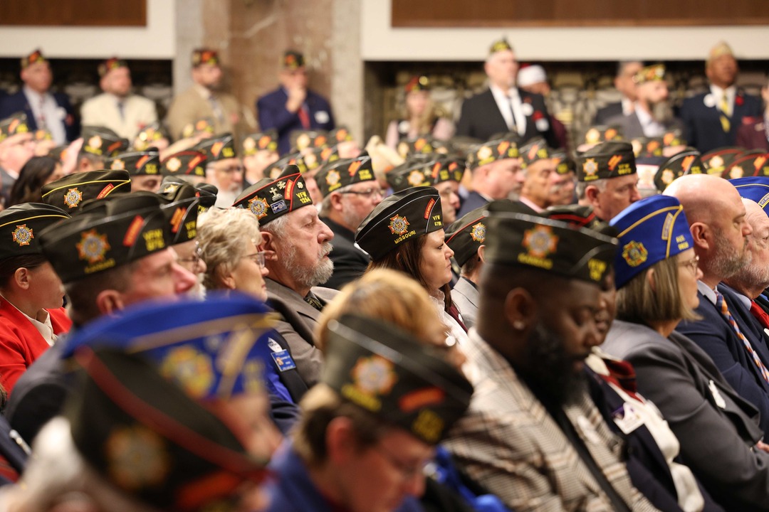 Una gran audiencia de veteranos con gorras de la VFW escucha atentamente durante un programa o evento.