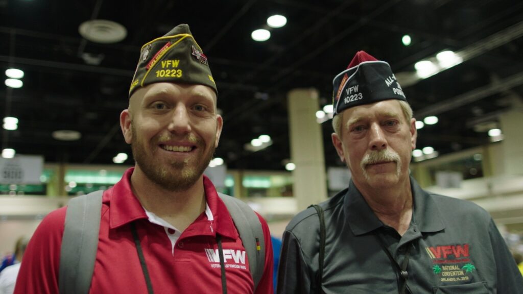 Two men wearing VFW hats smile at the camera, representing camaraderie at a veterans' event.