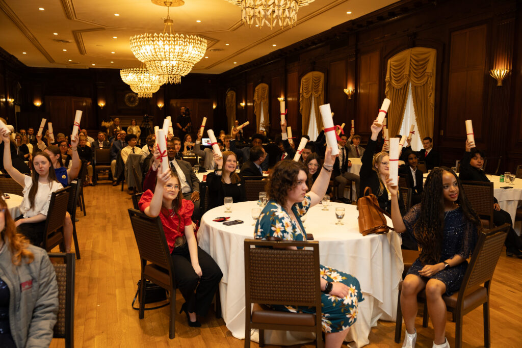 Students celebrate a ceremony, holding scrolls with red ribbons in a formal event setting.
