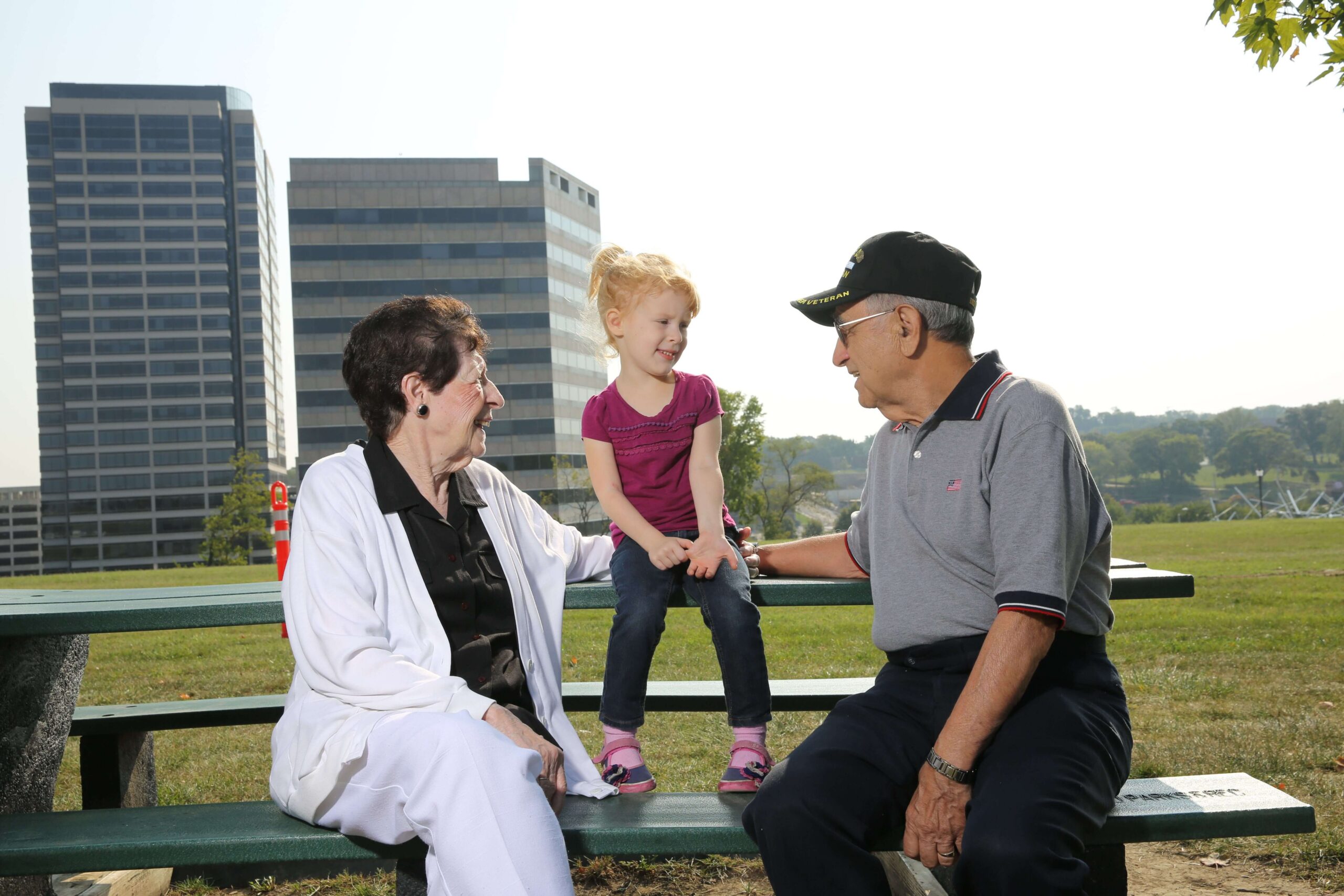 A smiling elderly woman and man engage with a young girl on a park bench, highlighting intergenerational connection.