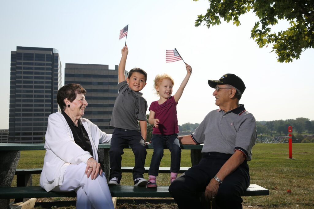 Two children joyfully wave small American flags while sitting with two smiling seniors at a park.