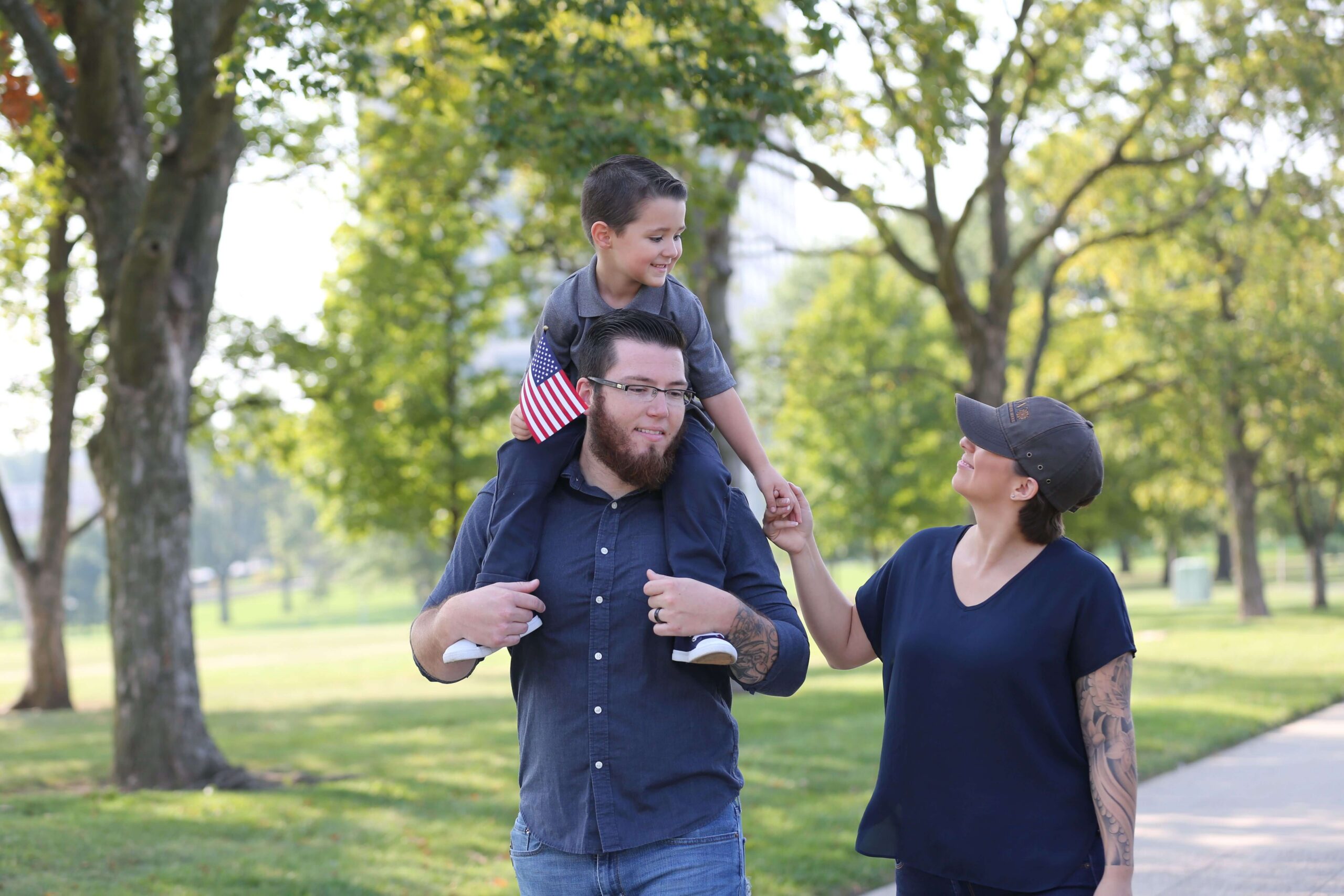 Family enjoying a walk in the park, with a boy on his father's shoulders holding a small American flag.