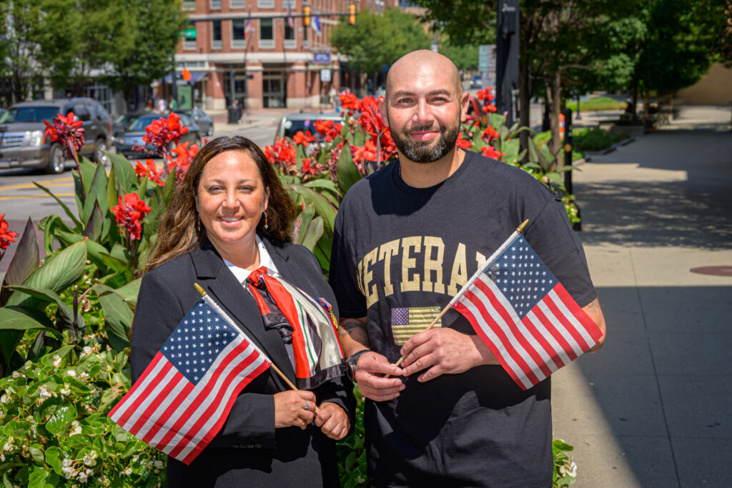 Two individuals, one in a suit and the other in a veteran t-shirt, hold American flags in front of vibrant flowers.