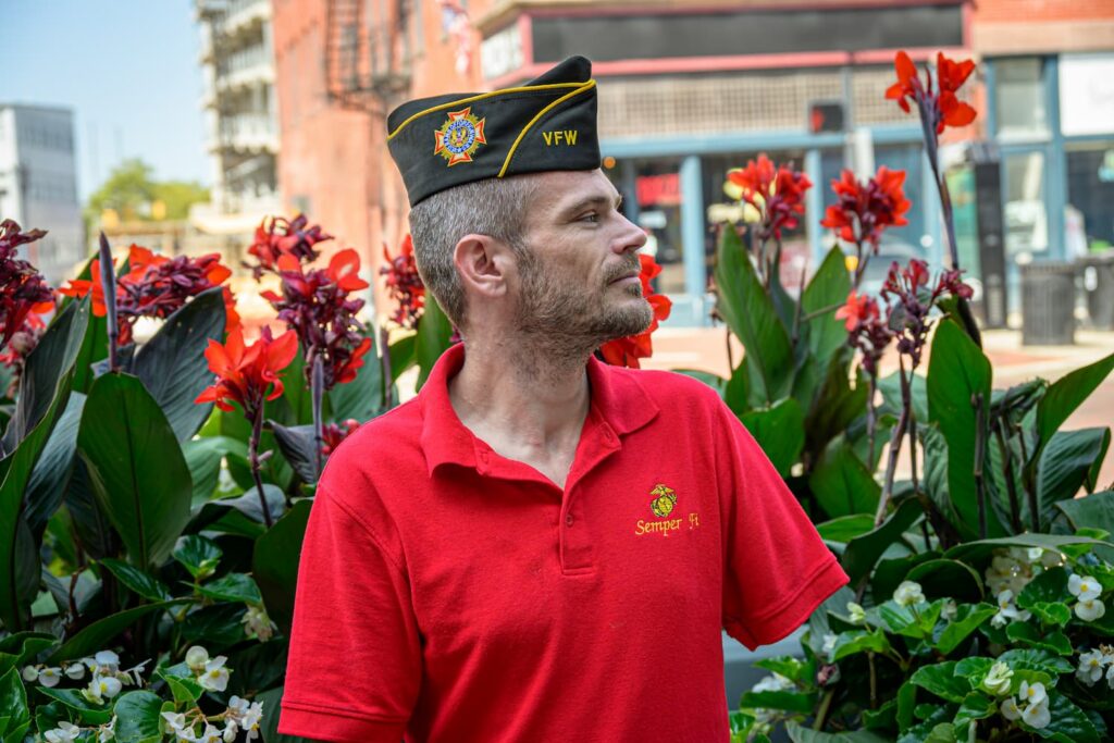 Veteran in a VFW hat stands proudly among vibrant flowers, symbolizing community engagement and educational outreach.