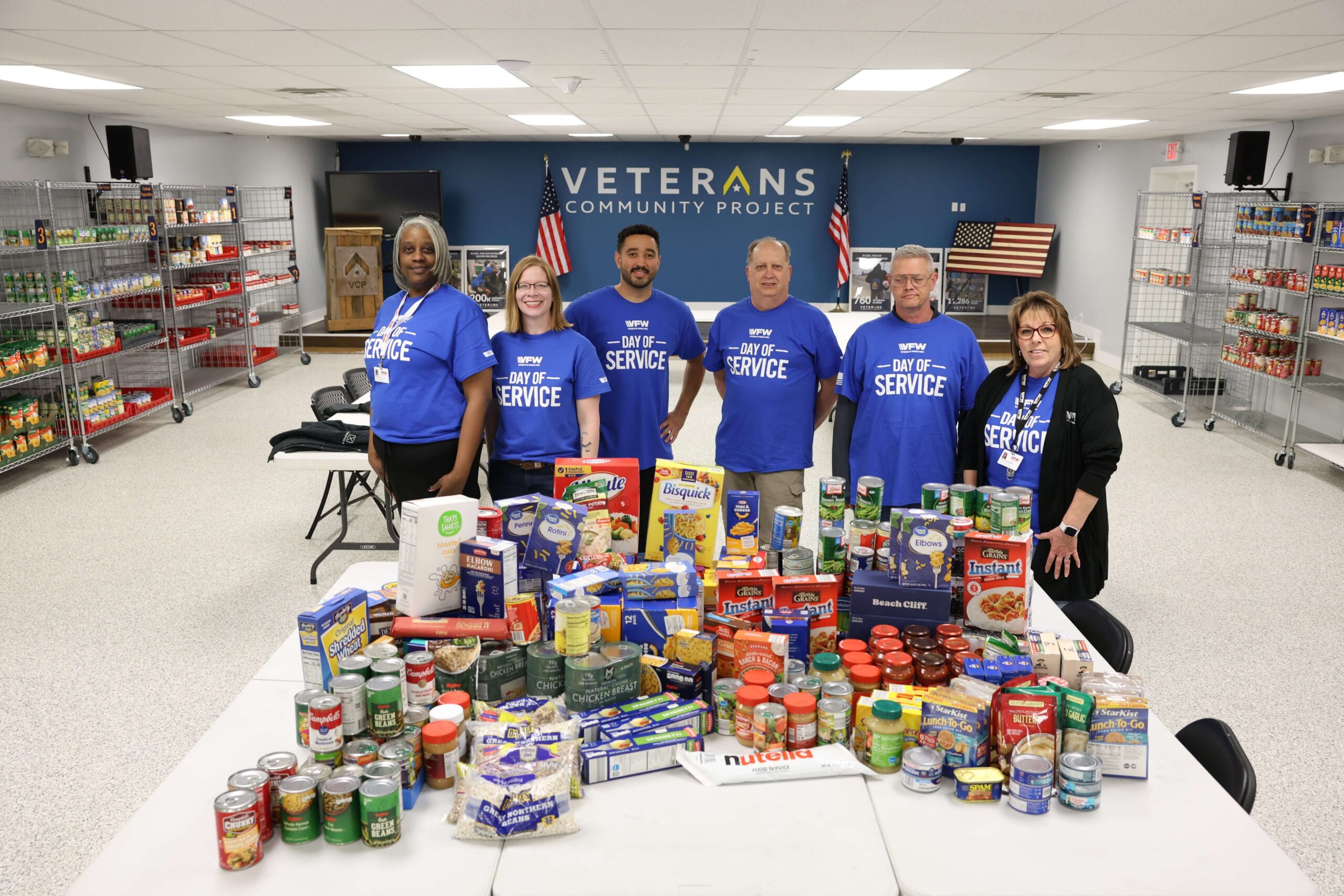 Six individuals wearing blue day of service shirts standing in front of a table with donated foot items and other assorted essentials.