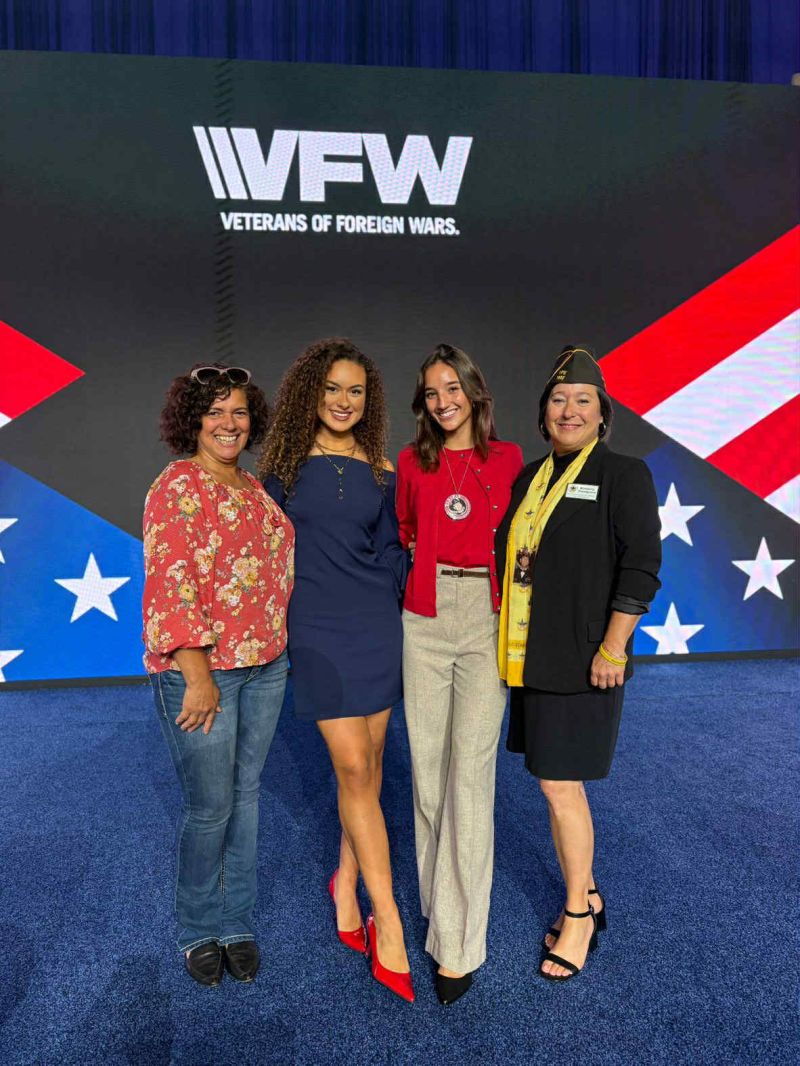 Four women pose together at a VFW convention, showcasing camaraderie against a backdrop featuring the VFW logo and patriotic symbols.