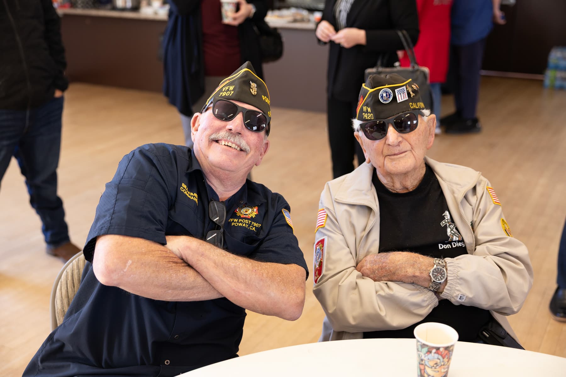 Two smiling veterans wearing sunglasses and hats sit at a table, showcasing camaraderie in a community gathering.