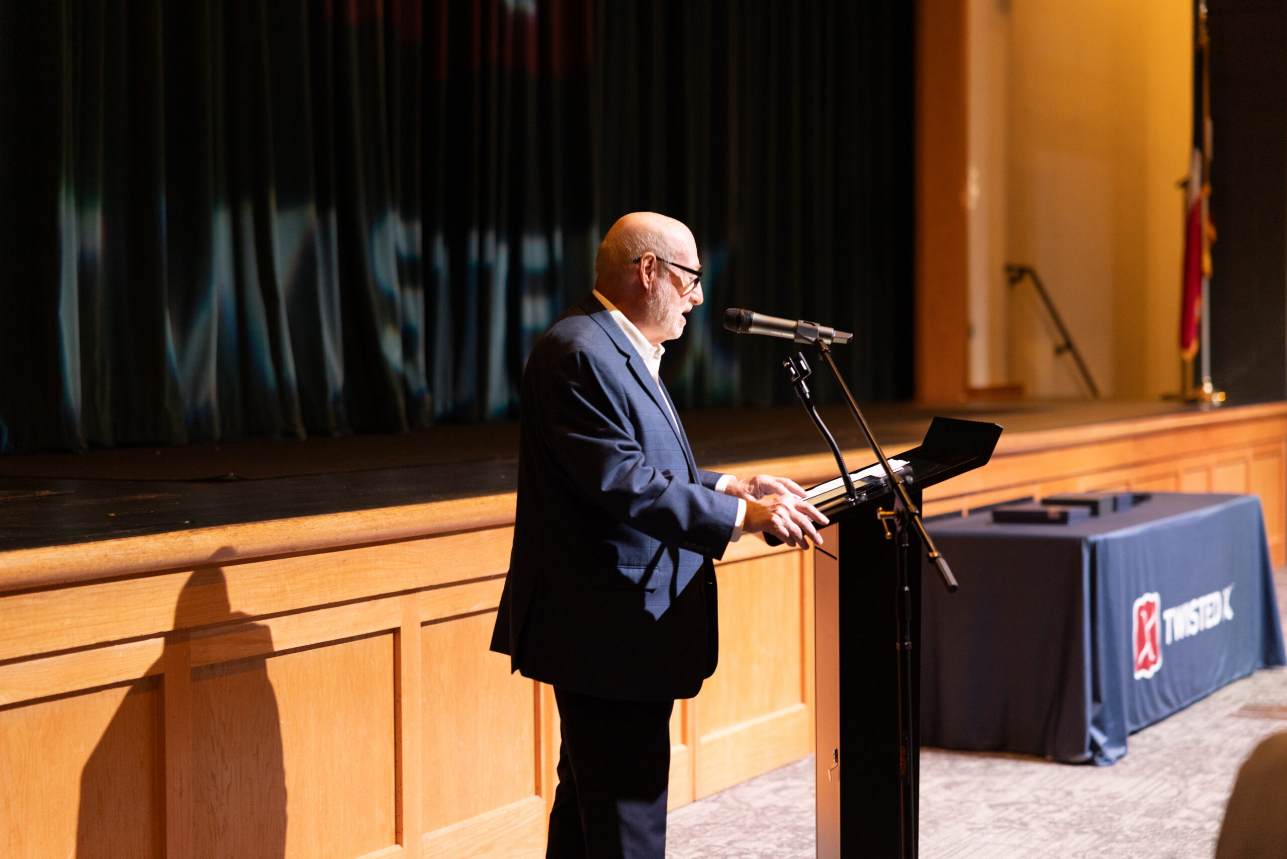A speaker addresses an audience from a podium on stage, with a table displaying event materials in the background.