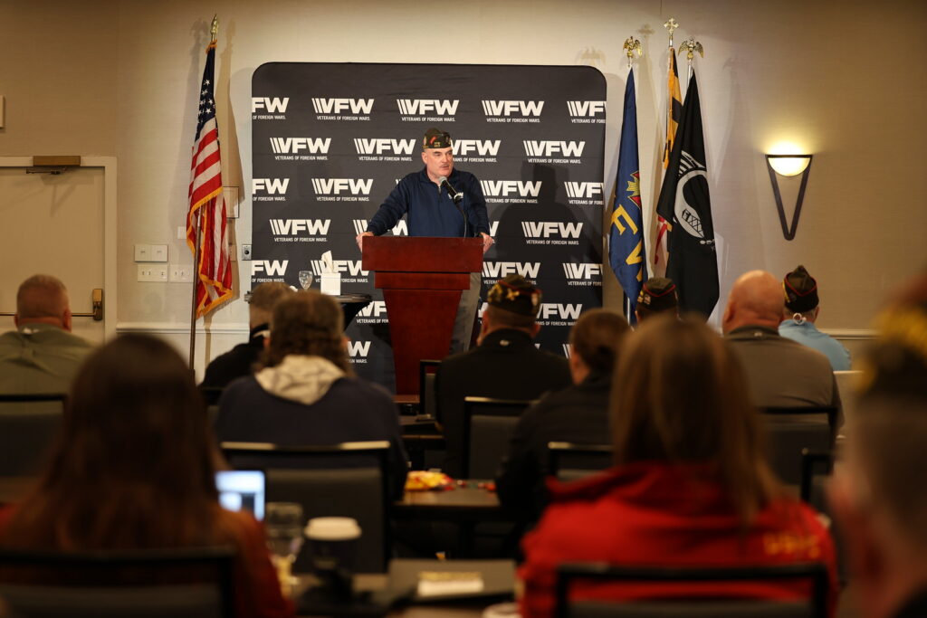 A speaker addresses an audience at a VFW event, with flags and a branded backdrop visible behind them.