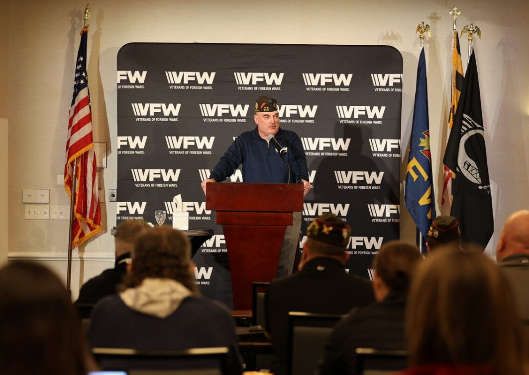 A speaker addresses an audience at a VFW event, with flags and a branded backdrop visible behind them.