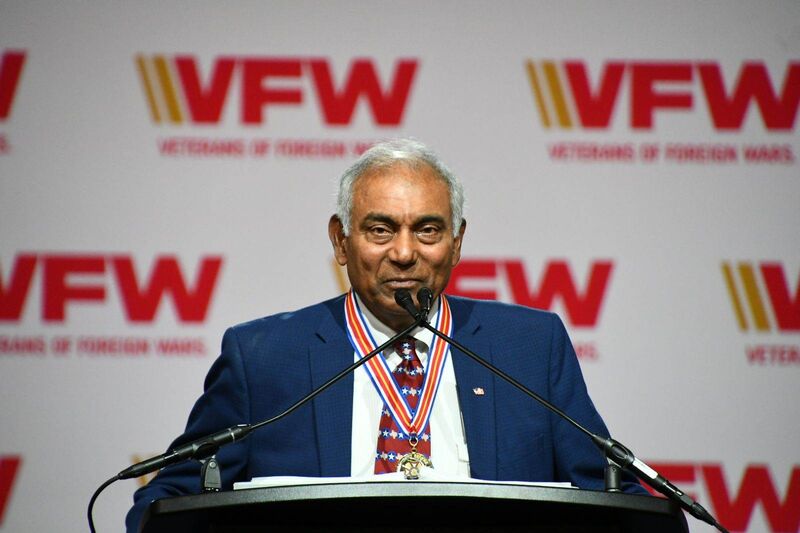 A speaker wearing a medal addresses an audience at a VFW event, promoting the organization's mission and values.