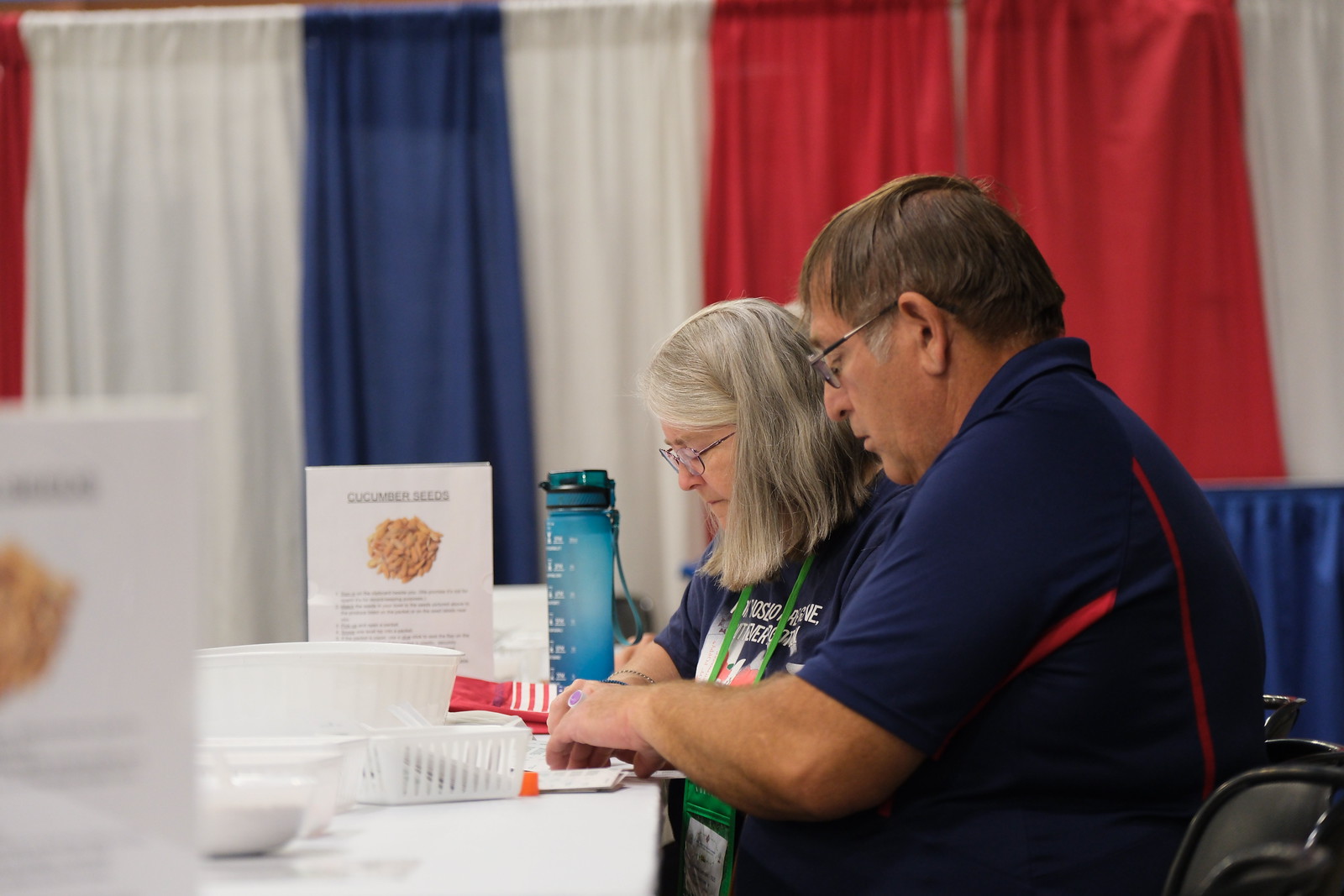 Two volunteers work at a table labeled with "Cucumber Seeds," focusing on tasks in a colorful event setting.