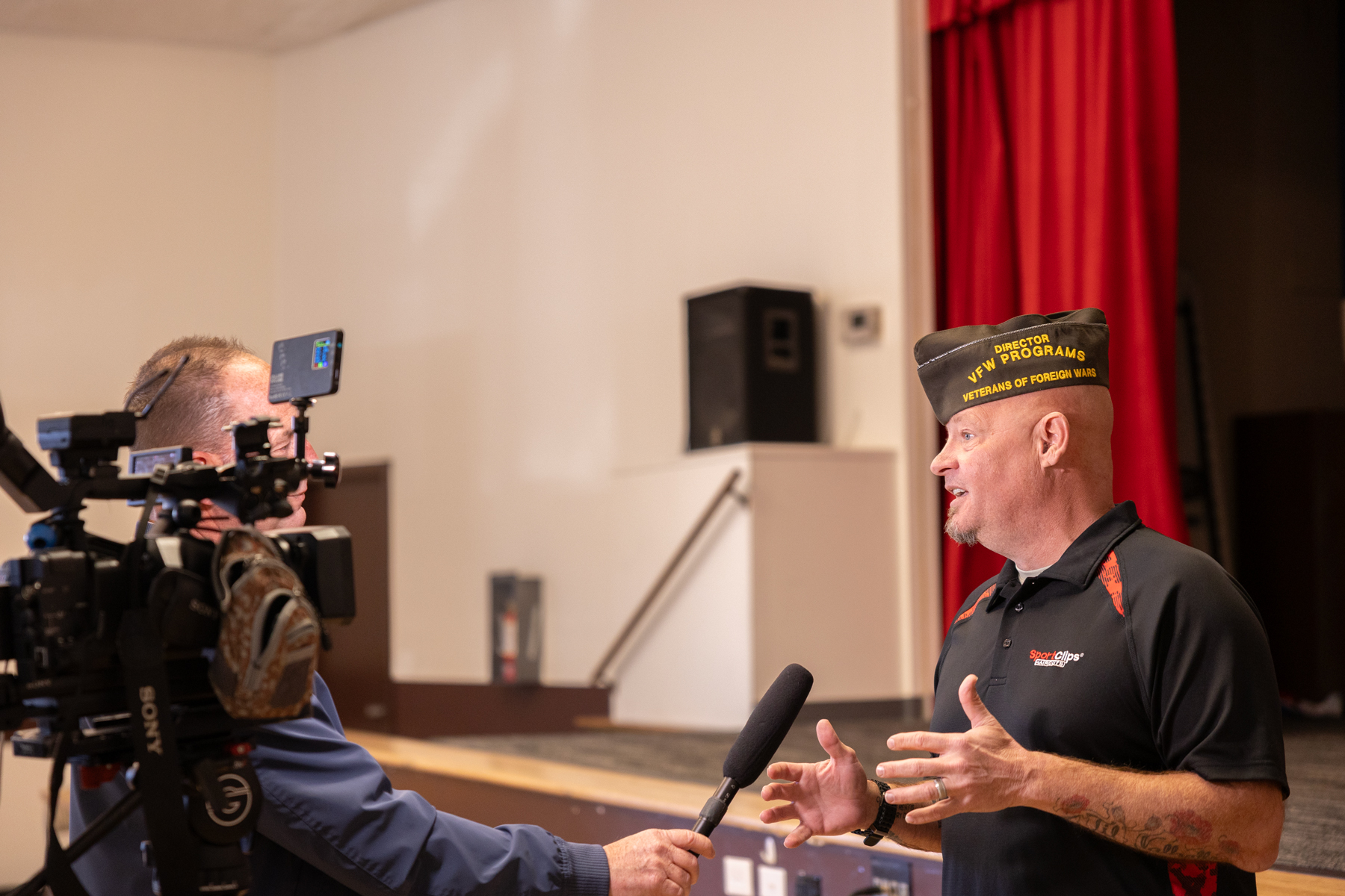 A veteran discusses community impact during an interview in a hall with red curtains and a camera crew present.
