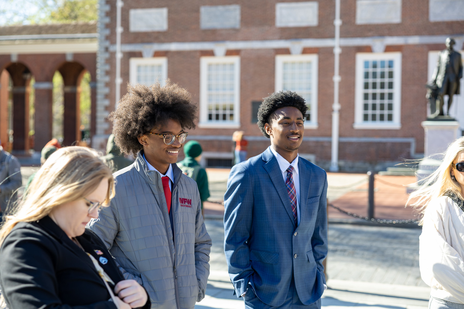 Two young men in formal attire smile and walk together outdoors, with a woman nearby and historical buildings in the background.